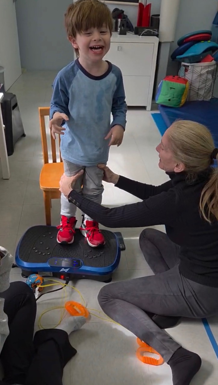 Dr. Tara Liddle supporting a smiling boy as he stands on a vibration plate during a physical therapy session.