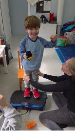 Dr. Tara Liddle supporting a smiling boy as he stands on a vibration plate.