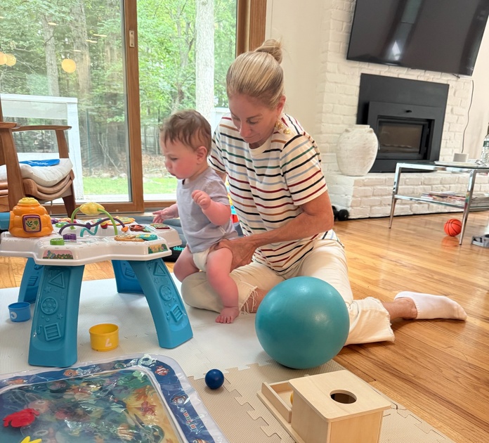 A pediatric physical therapist supporting a baby standing at an activity table during a home therapy session.