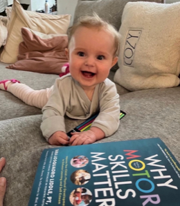 A smiling baby propped up on a couch beside a copy of Why Motor Skills Matter.